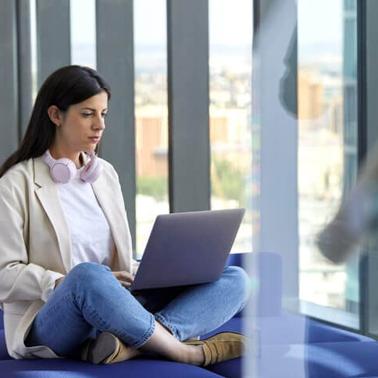 A woman sits comfortably working on her laptop in a modern office setting with large windows.