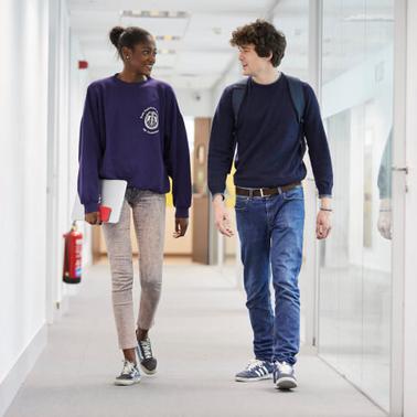 Two young adults, a male and a female, are walking and talking in a brightly lit corridor.