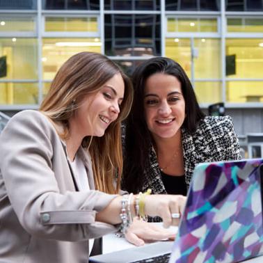 Two women are smiling and working together on a laptop outdoors.