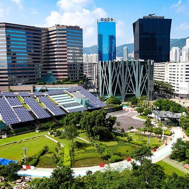 A modern urban park with solar panels and lush greenery surrounded by tall commercial buildings and a clear blue sky.
