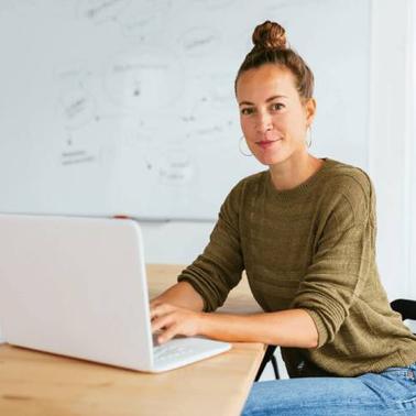A woman sitting at a desk in an office, working on a laptop with a whiteboard in the background.