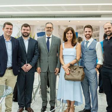 A group of six adults, three men and three women, pose for a photograph at an indoor event with other attendees in the background.
