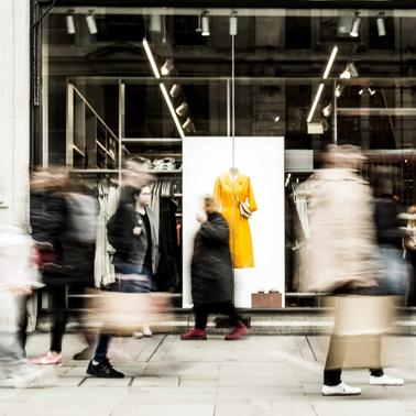 Blurred figures walking past a fashion store display with a bright yellow jacket on a mannequin.
