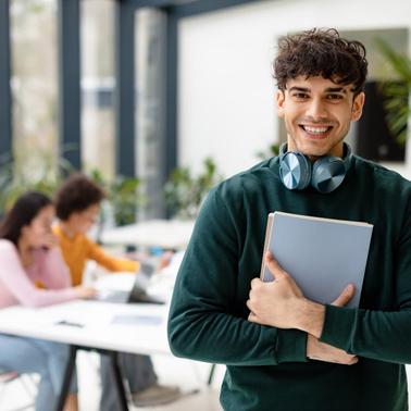 A young man with headphones around his neck holding a laptop smiles at the camera, with people in the background focusing on their work in a bright, modern workspace.