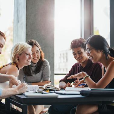 A group of five young adults happily studying together at a table in a sunny room.