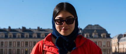 A woman in a red jacket stands in front of a historical building on a sunny day.
