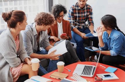 A group of five diverse people engaging in a discussion with digital devices and papers on a round table.