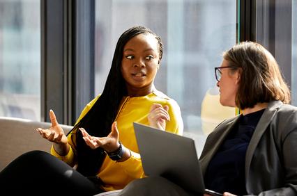 Two women are engaged in a conversation, one gesturing while the other listens attentively.