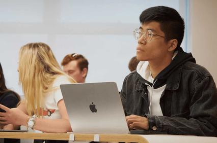 A young man with glasses and a denim jacket sits at a desk with a laptop in a classroom, looking to the side while other students are in the background.