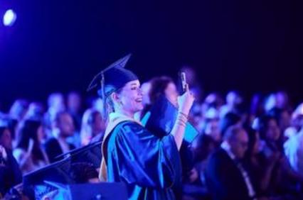 A graduate smiles while taking a selfie during a graduation ceremony.