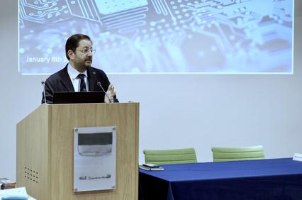 A man in formal attire speaks at a podium in front of a technology-themed backdrop.
