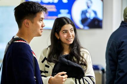 A young man and woman are smiling and engaging in a conversation in a busy indoor setting.