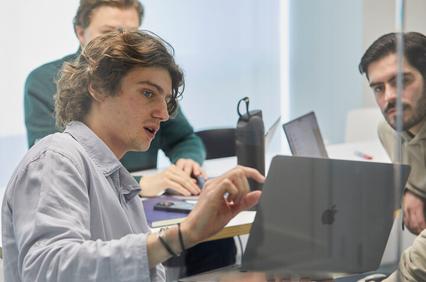 A group of young men are engaged in a collaborative discussion while reviewing work on a laptop.