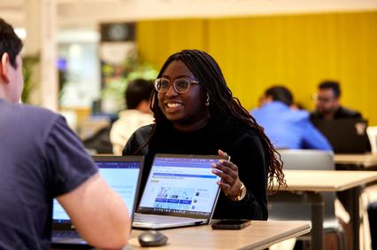 A woman with glasses discussing over a laptop in a busy office area.