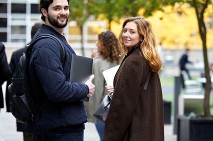 Two young professionals smiling and looking back while walking in a city setting with laptops in hand.