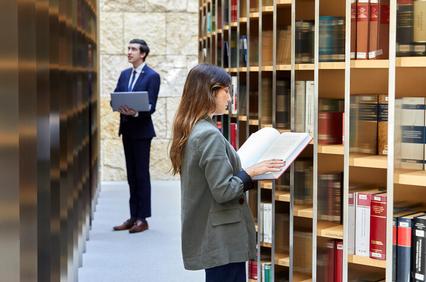 A woman and a man, dressed in business attire, are reading books in a library with floor-to-ceiling shelves filled with books.