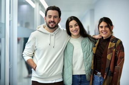 Three young adults, two women and one man, smiling and posing together indoors.