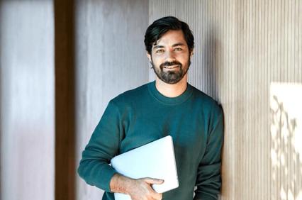 A man with a beard smiling and holding a laptop in a modern office environment.