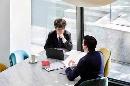 Two men in business attire having a discussion at a modern office table with a laptop and documents in front of them.