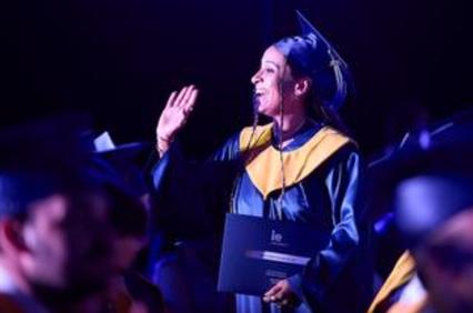 A graduate joyfully waves while holding their diploma during a graduation ceremony.