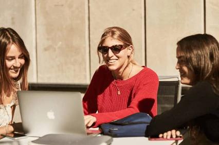 Three women sit at a table, smiling and talking with a laptop open in front of them.