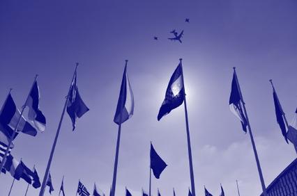 A monochrome image of multiple flags on poles against a sky with an airplane flying overhead.
