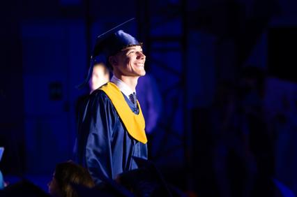 A smiling graduate in a blue cap and gown stands proudly under stage lights.