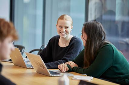 Two women are engaged in a lively conversation at a table with laptops.