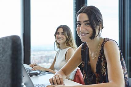 Two women are sitting at a desk in an office with a cityscape view.