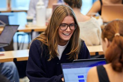 A smiling young woman with glasses works on a laptop in a classroom setting.