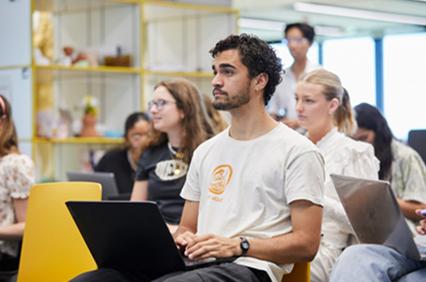 A diverse group of people are attending a seminar, with a focus on a man in a white t-shirt sitting in the front.