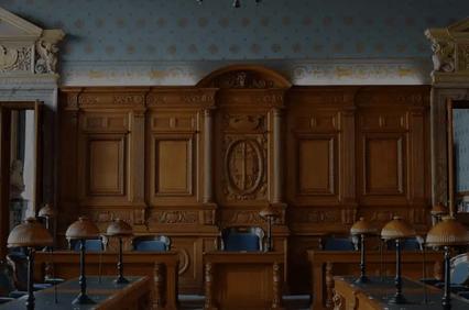 A panoramic view of a grand, classically designed courtroom with wooden furniture and large, ornate windows.