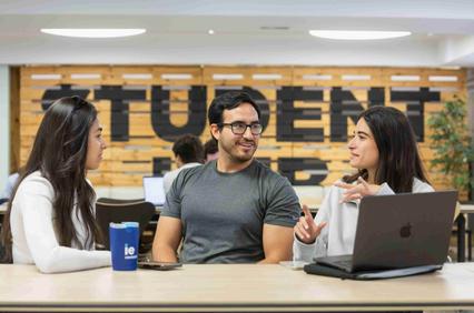Three students are engaged in a discussion around a table in a study area.