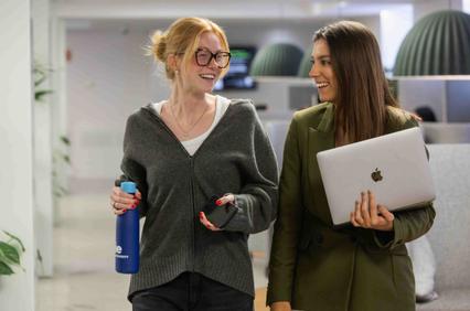 Two women are walking and smiling while engaging in conversation in a modern office setting.