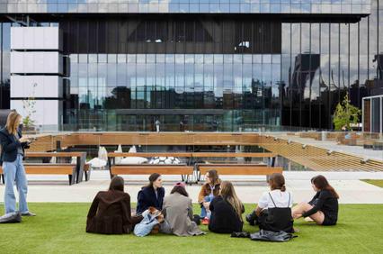 A group of young people are sitting on a lawn in front of a modern building talking and sharing.