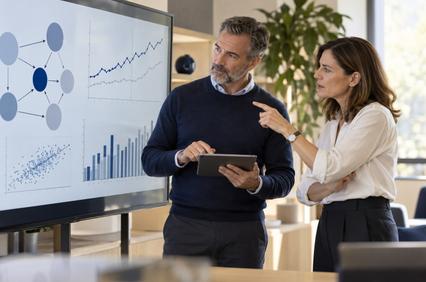 A man and a woman are discussing data analysis while looking at a digital screen displaying various graphs and charts.