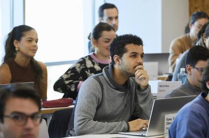 A diverse group of students attentively listening in a classroom setting, with laptops open and some taking notes.