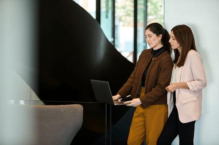 Two women standing and looking at a laptop in a modern office setting.