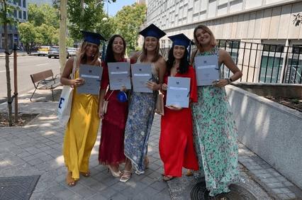 A group of five young women in graduation attire holding diplomas on a city street.