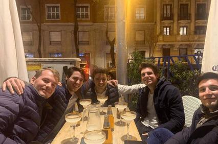A group of five young men smiling at a table with drinks in an outdoor dining area at night.