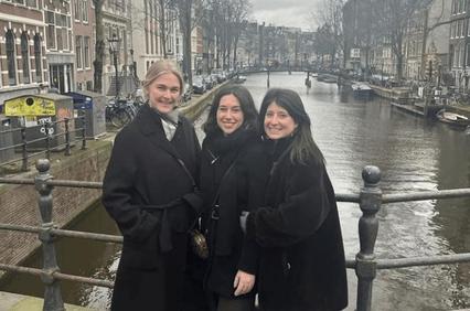 Three women smiling in front of a canal in an urban setting.