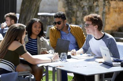 A group of young adults engaging in a discussion at an outdoor café table, with laptops and smartphones visible.