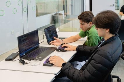 Two students are collaborating on coding tasks at a table with laptops.
