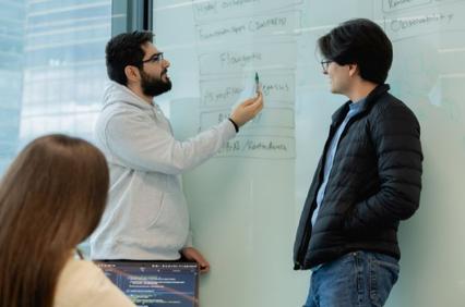 Two men are engaged in a discussion while reviewing notes on a whiteboard.