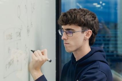 A young man is writing on a glass board while deep in thought.