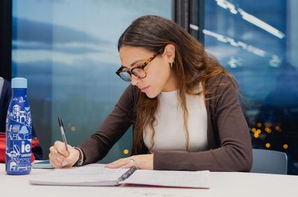 A young woman is studying and taking notes at a table with a water bottle beside her.