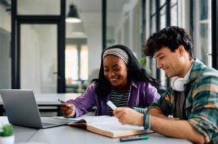 Two students are studying together at a table with a laptop and books.