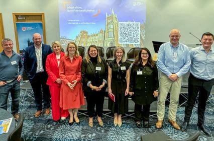A group of nine individuals posing for a photo at an event, with a presentation screen displaying a QR code and the name 'School of Law, Queen's University Belfast' in the background.