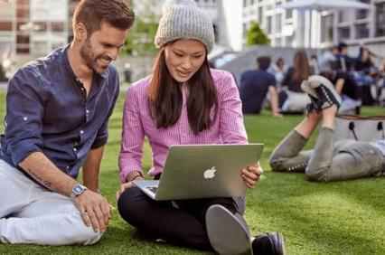 A man and a woman are sitting on the grass, looking at a laptop together with other people in the background.