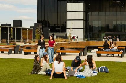 A group of young people sitting and standing on a grassy area at a modern university campus.